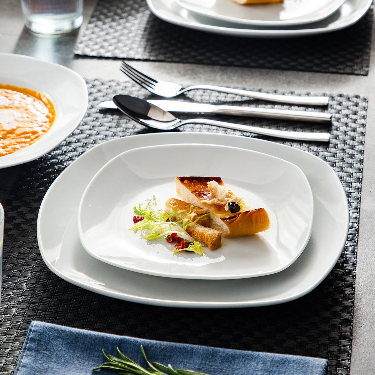 White square dinnerware with a plated appetizer and silverware on a black woven placemat