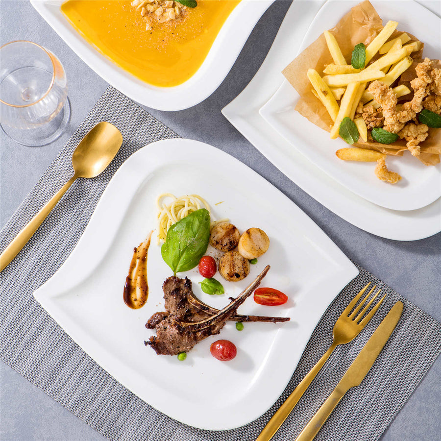 Overhead formal table setting with white square plates of grilled lamb chops, soup, fried chicken, and fries beside gold flatware