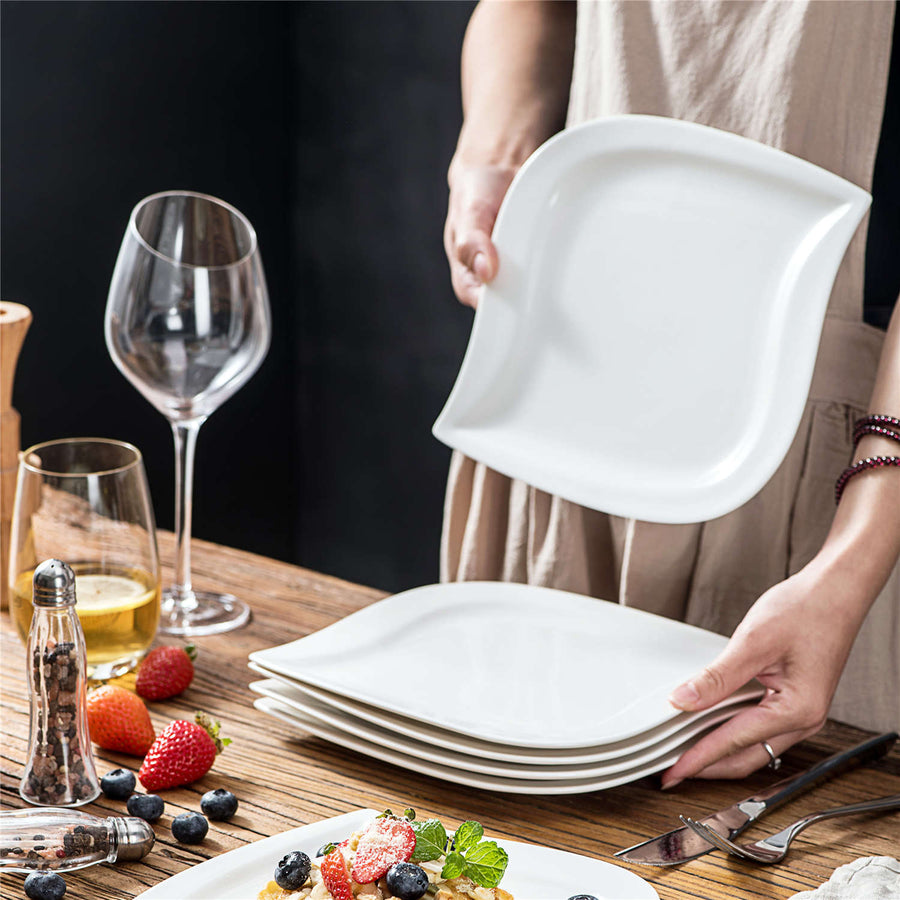 Hands holding elegant white wavy dinner plates, with more stacked below, on a rustic wooden table set with fruit and drinks